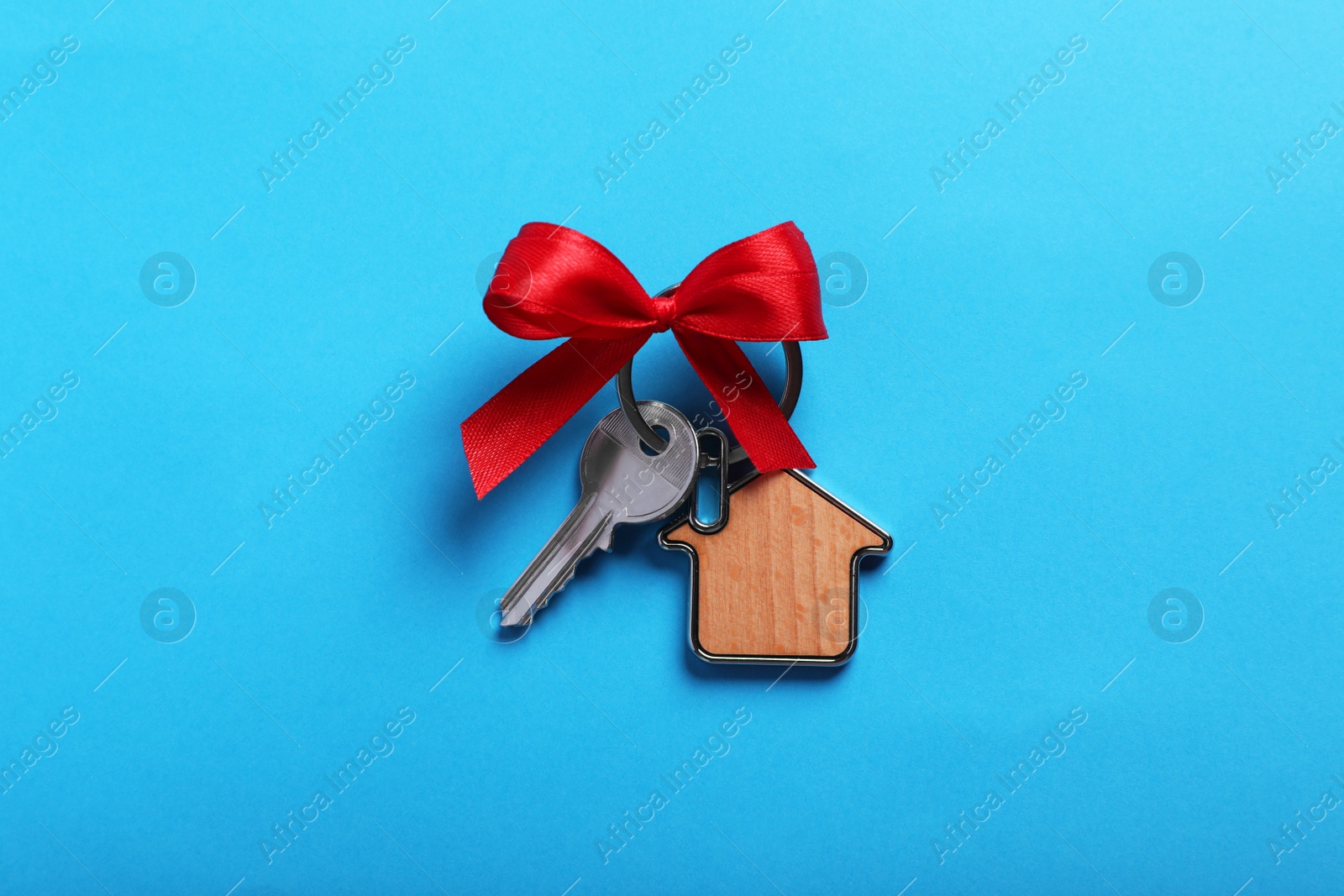 Photo of Key with trinket in shape of house and bow on light blue background, top view. Housewarming party