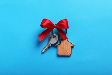 Key with trinket in shape of house and bow on light blue background, top view. Housewarming party