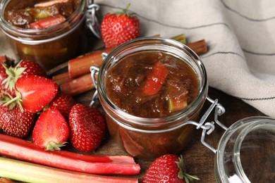 Photo of Jars of tasty rhubarb jam, fresh stems and strawberries on wooden table