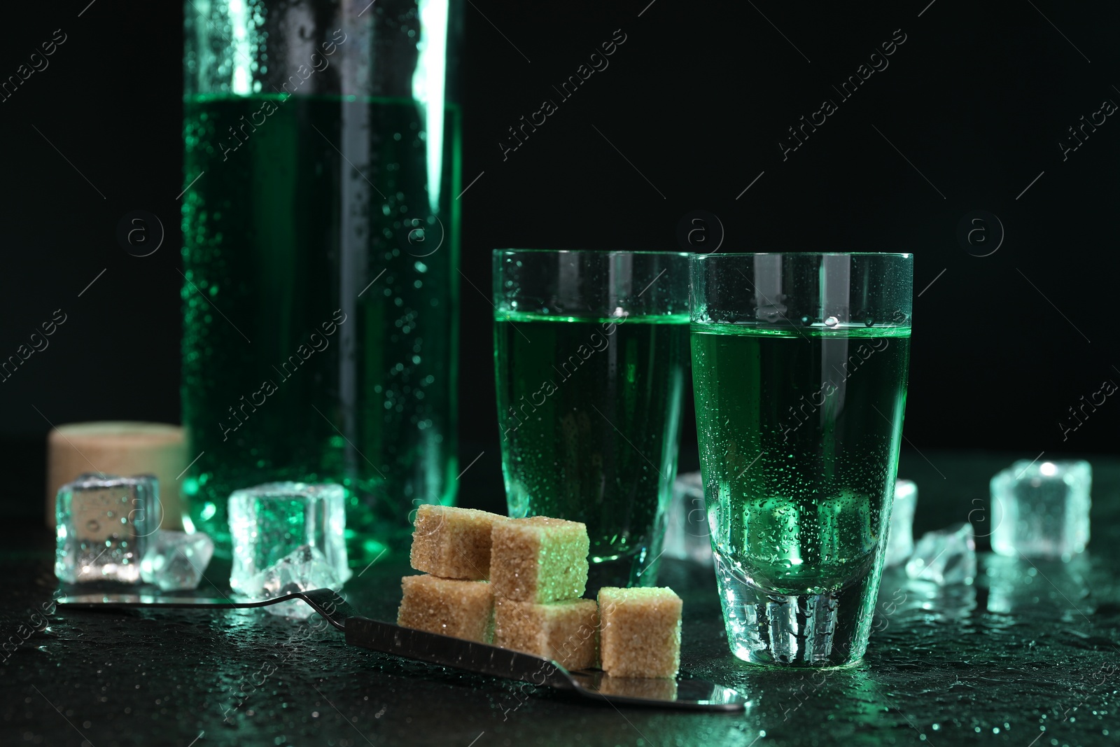 Photo of Absinthe in shot glasses, spoon, brown sugar and ice cubes on gray table against dark background. Alcoholic drink