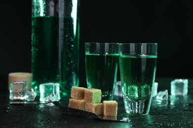 Photo of Absinthe in shot glasses, spoon, brown sugar and ice cubes on gray table against dark background. Alcoholic drink
