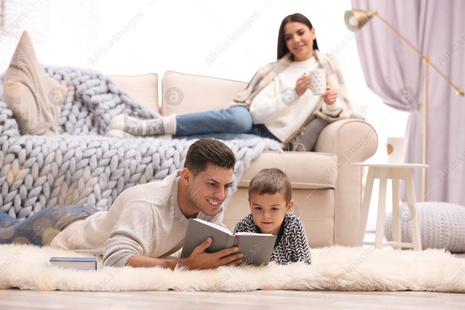 Photo of Father and his son reading books while mother resting on sofa at home. Winter vacation