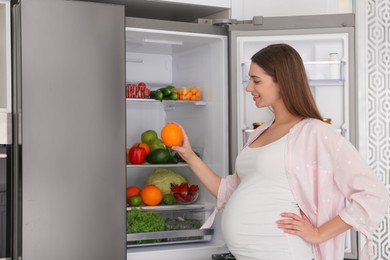Photo of Young pregnant woman with orange near fridge at home. Healthy eating