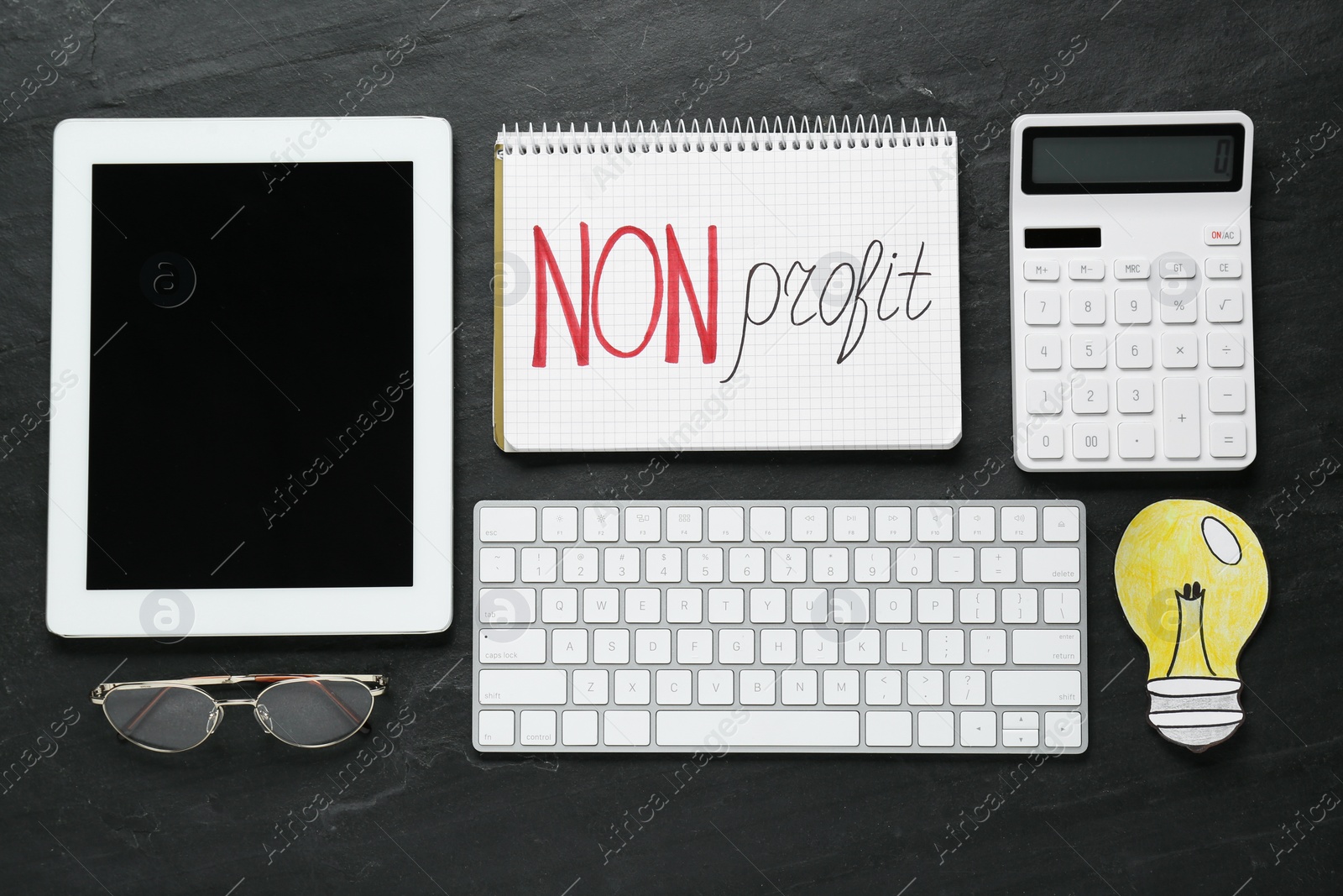 Photo of Notebook with phrase Non Profit, calculator, computer keyboard, tablet and glasses on black table, flat lay