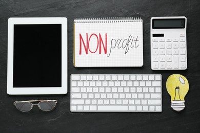 Photo of Notebook with phrase Non Profit, calculator, computer keyboard, tablet and glasses on black table, flat lay