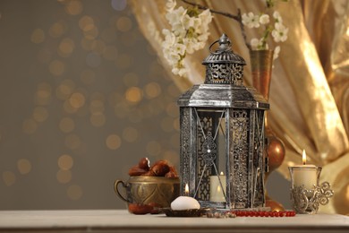 Photo of Arabic lantern, misbaha, candles, dates and flowers on table against blurred lights, space for text