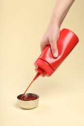 Woman squeezing tasty ketchup from bottle into bowl on beige background, closeup