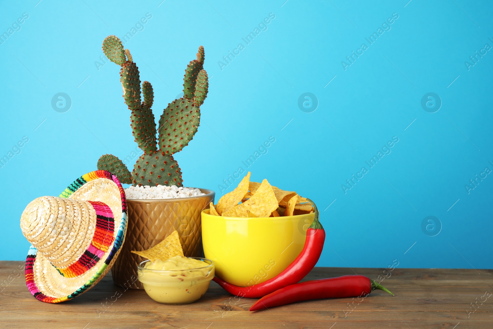 Photo of Mexican sombrero hat, cactus, chili peppers, nachos chips and guacamole in bowls on wooden table against light blue background, space for text