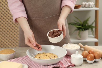 Photo of Cooking sweet cookies. Woman adding chocolate chips to dough at table in kitchen, closeup