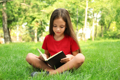 Photo of Cute little girl reading book on green grass in park