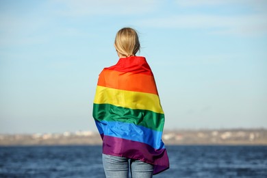 Woman wrapped in bright LGBT flag near river, back view