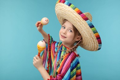 Photo of Cute girl in Mexican sombrero hat and poncho dancing with maracas on light blue background