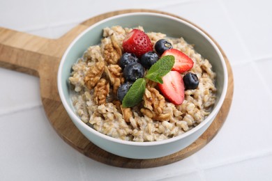 Photo of Tasty oatmeal with strawberries, blueberries and walnuts in bowl on white tiled table, closeup