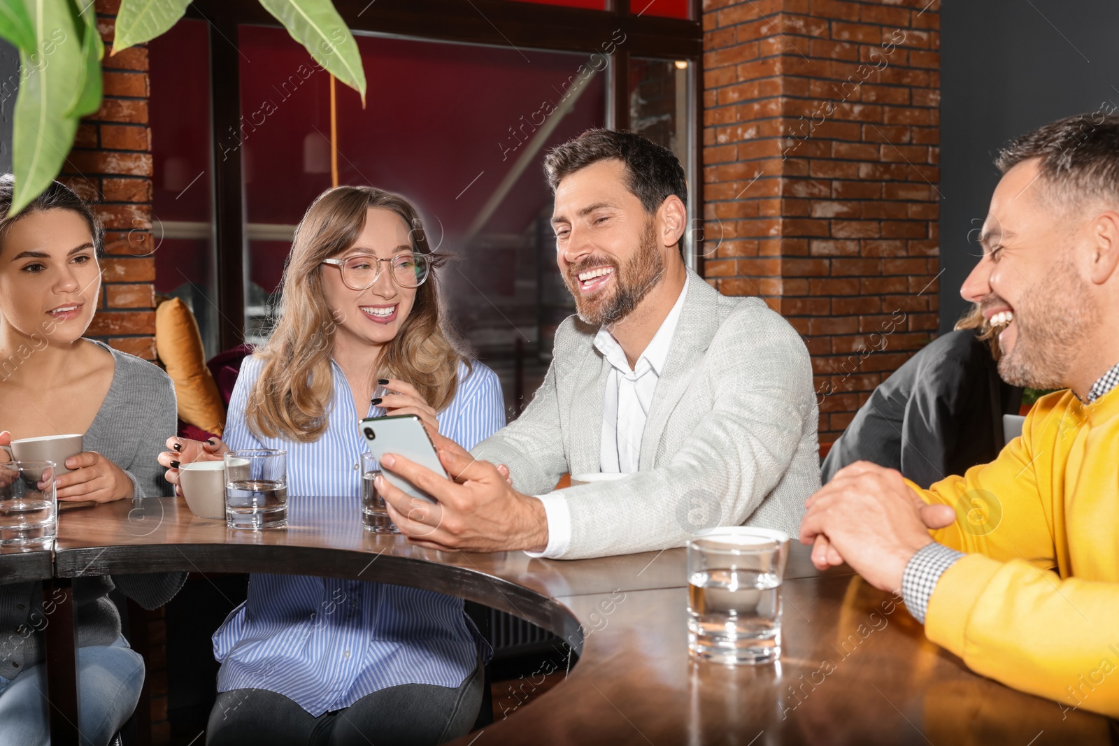 Photo of Handsome man showing something funny in smartphone to his friends in cafe