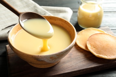 Photo of Spoon of pouring condensed milk over bowl on table, space for text. Dairy products