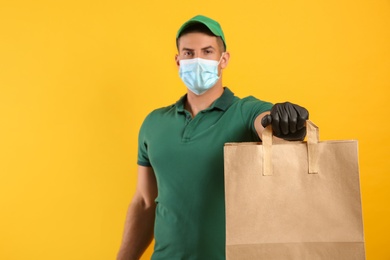 Photo of Courier in medical mask holding paper bag with takeaway food on yellow background, space for text. Delivery service during quarantine due to Covid-19 outbreak