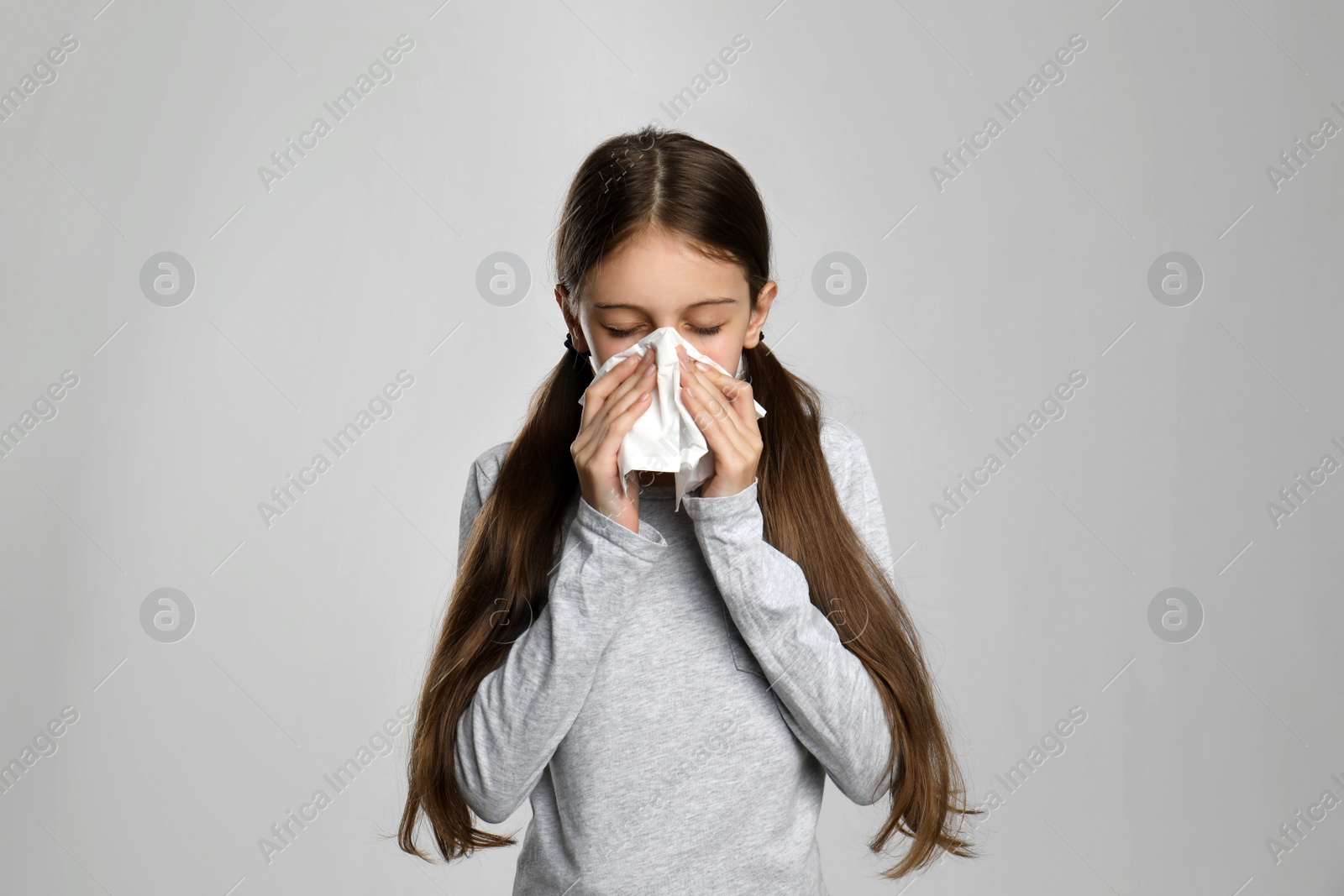 Photo of Little girl blowing nose into paper tissue on light grey background. Seasonal allergy