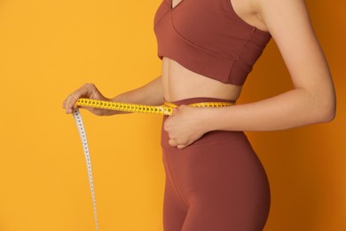 Photo of Woman in sportswear measuring waist with tape on yellow background, closeup