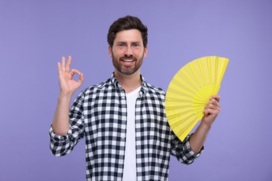Photo of Happy man holding hand fan and showing ok gesture on purple background