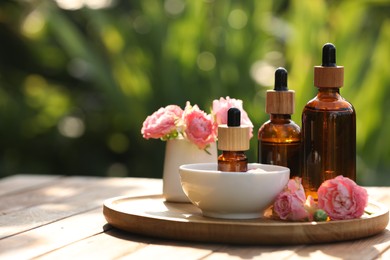 Photo of Bottles of rose essential oil and flowers on wooden table outdoors, space for text