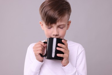Cute boy drinking beverage from black ceramic mug on light grey background