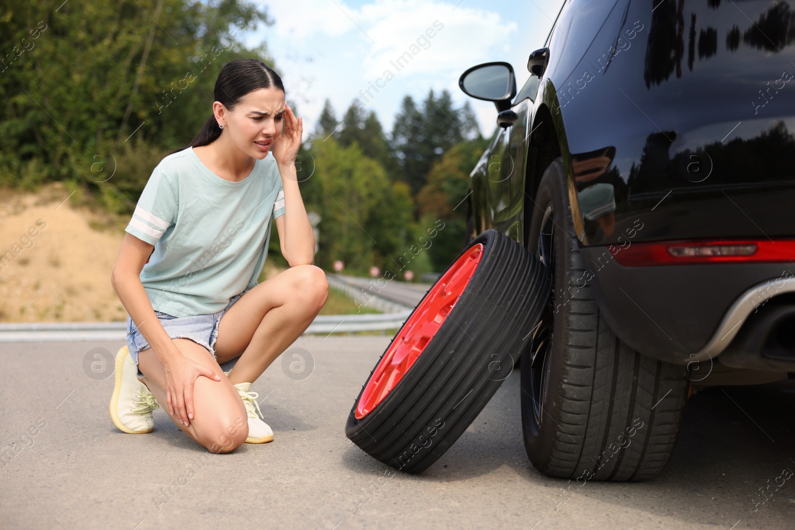 Photo of Tire puncture. Stressed woman with new wheel near car on roadside outdoors