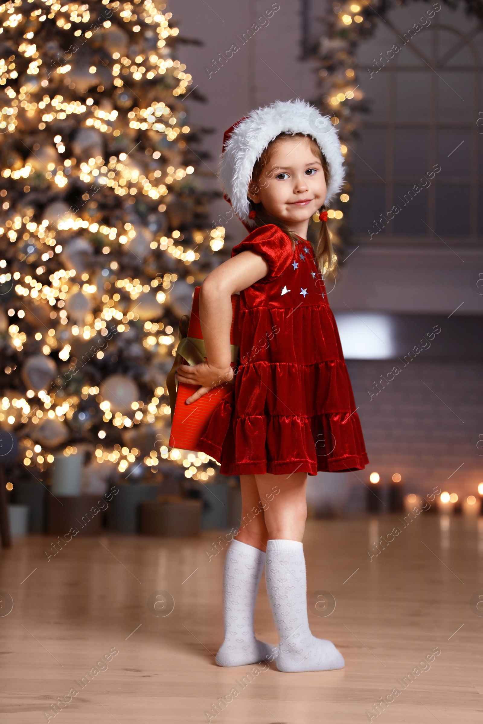 Photo of Cute little child wearing Santa hat hiding Christmas gift behind her back at home