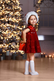 Cute little child wearing Santa hat hiding Christmas gift behind her back at home