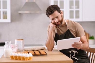 Photo of Man with freshly baked cookies watching online cooking course via tablet in kitchen