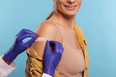 Nurse sticking adhesive bandage on woman's arm after vaccination on light blue background, closeup