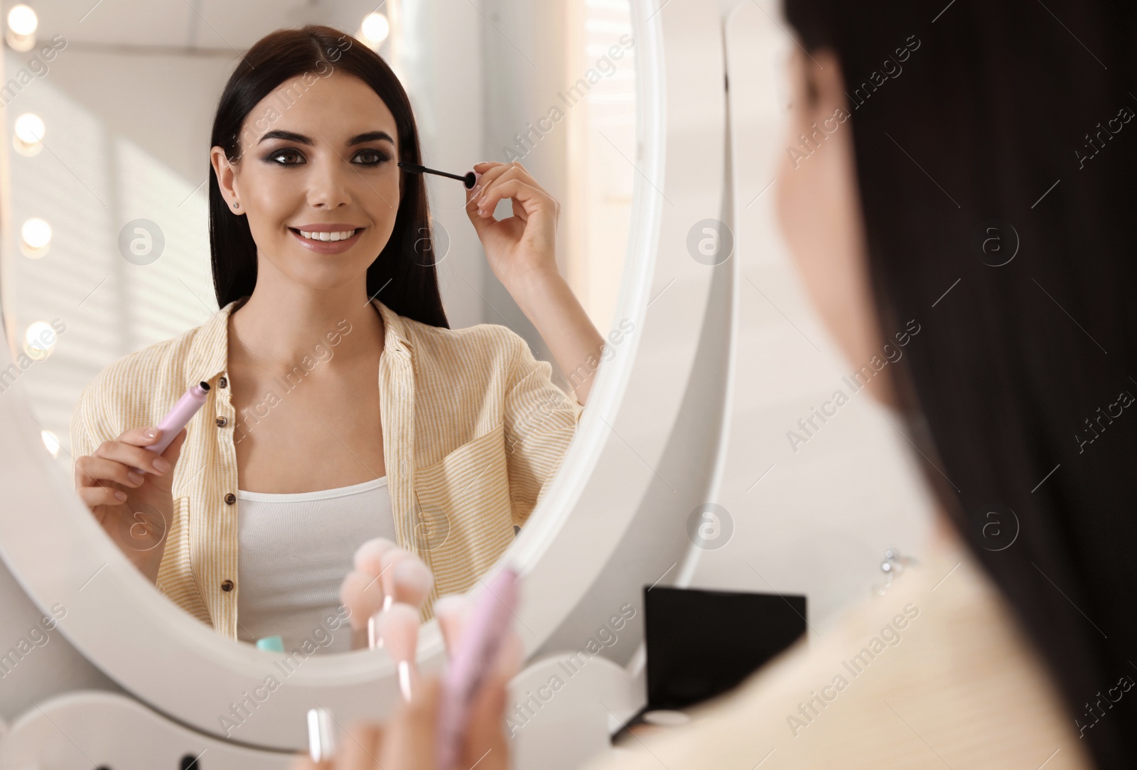 Photo of Beautiful young woman applying makeup near mirror indoors