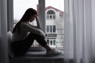 Photo of Unhappy young woman near window indoors, space for text. Loneliness concept