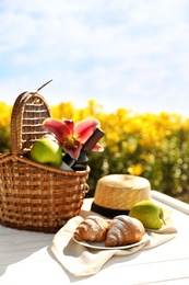 Photo of Picnic basket with bottle of wine and food on white wooden table in lily field