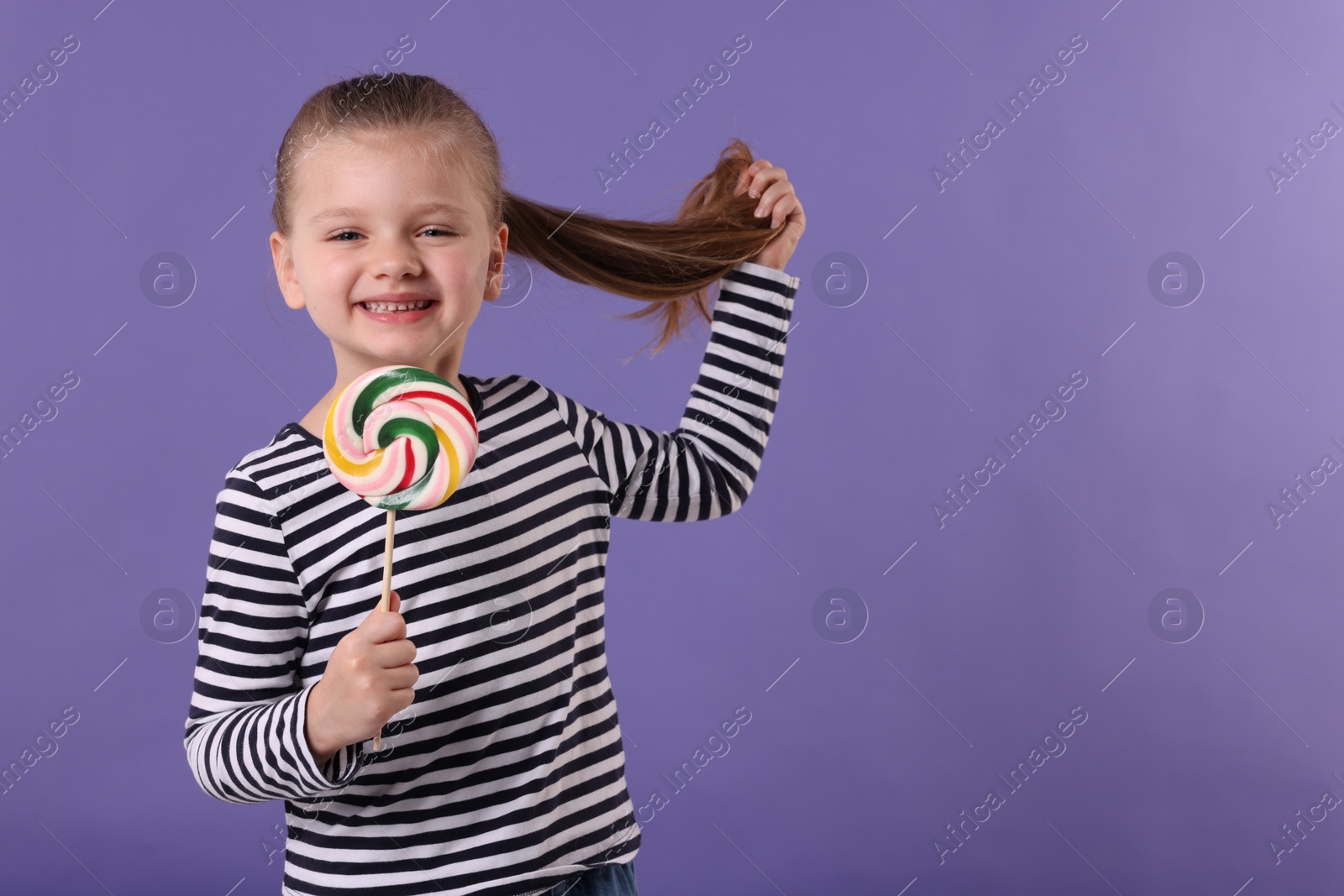 Photo of Happy little girl with colorful lollipop swirl on violet background, space for text