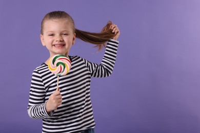 Photo of Happy little girl with colorful lollipop swirl on violet background, space for text