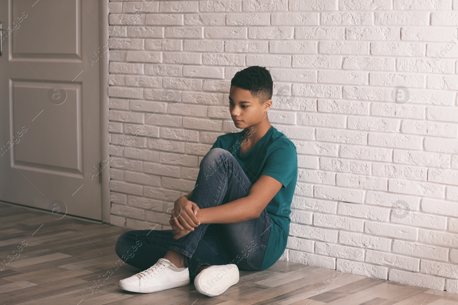 Photo of Upset African-American teenage boy sitting alone on floor near wall