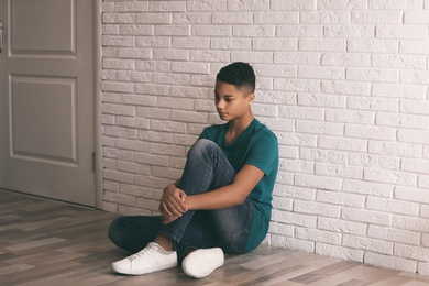 Upset African-American teenage boy sitting alone on floor near wall