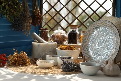 Photo of Many different dry herbs, flowers and plates on white wooden table near window