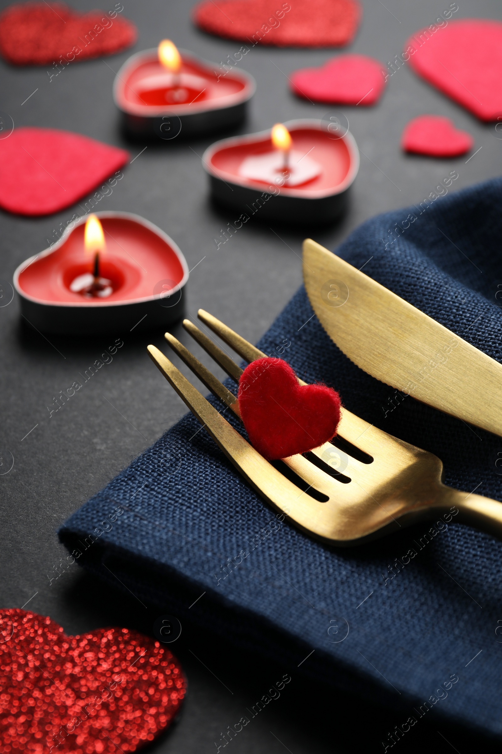 Photo of Cutlery set, burning candles and decorative hearts on black background, closeup. Romantic table setting