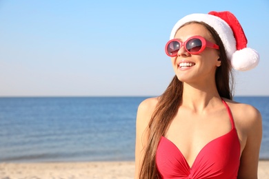 Photo of Beautiful young woman in Santa hat and bikini on beach, space for text. Christmas vacation