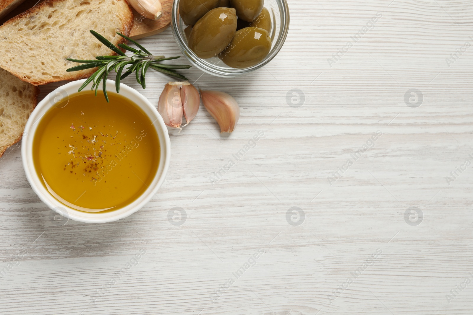 Photo of Bowl of fresh oil, bread, rosemary and garlic on white wooden table, flat lay. Space for text