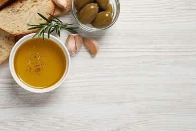 Photo of Bowl of fresh oil, bread, rosemary and garlic on white wooden table, flat lay. Space for text