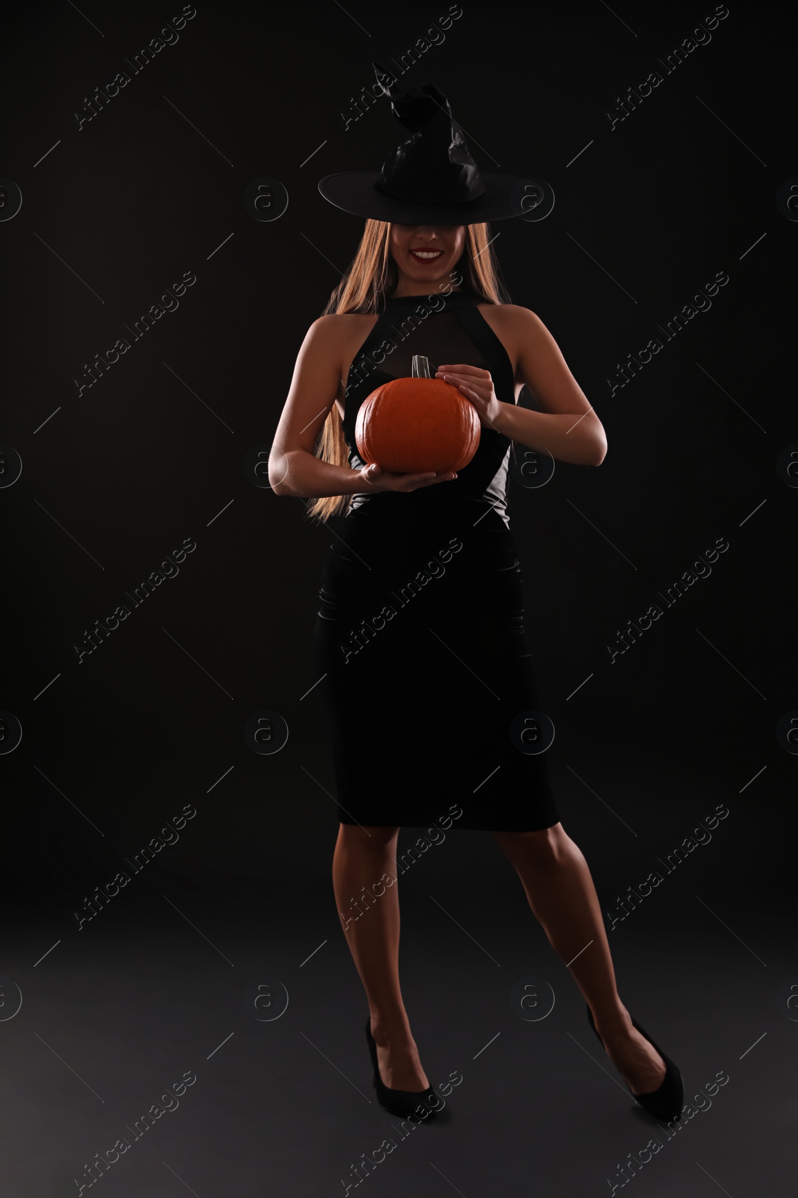 Photo of Young woman wearing witch costume with pumpkin on black background. Halloween party