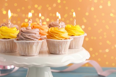 Photo of White stand with tasty birthday cupcakes on light blue wooden table against blurred lights, closeup