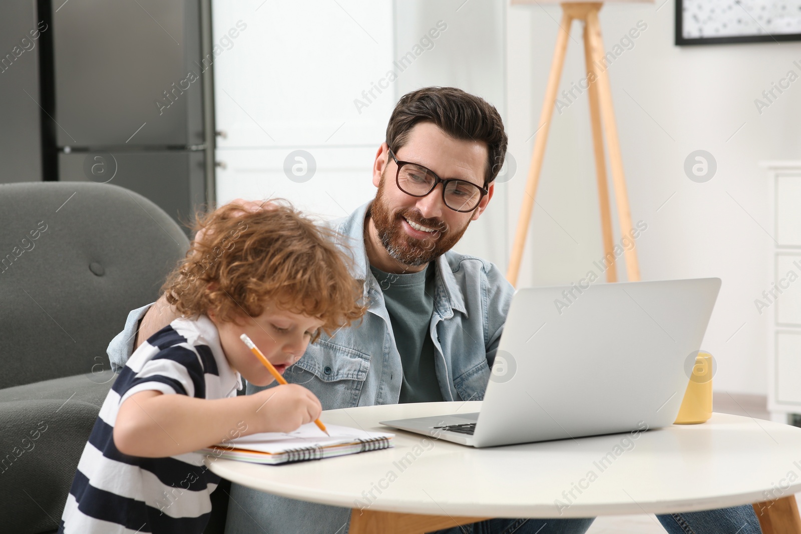 Photo of Man working remotely at home. Father using laptop while his son drawing at desk