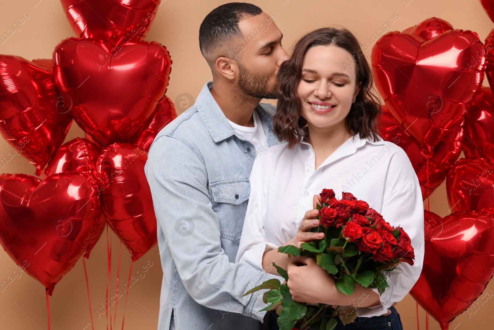 Photo of Happy couple celebrating Valentine's day. Beloved woman with bouquet of red roses near heart shaped air balloons on beige background