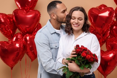 Photo of Happy couple celebrating Valentine's day. Beloved woman with bouquet of red roses near heart shaped air balloons on beige background