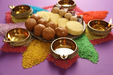 Photo of Diwali celebration. Tasty Indian sweets, diya lamps and colorful rangoli on violet table, closeup