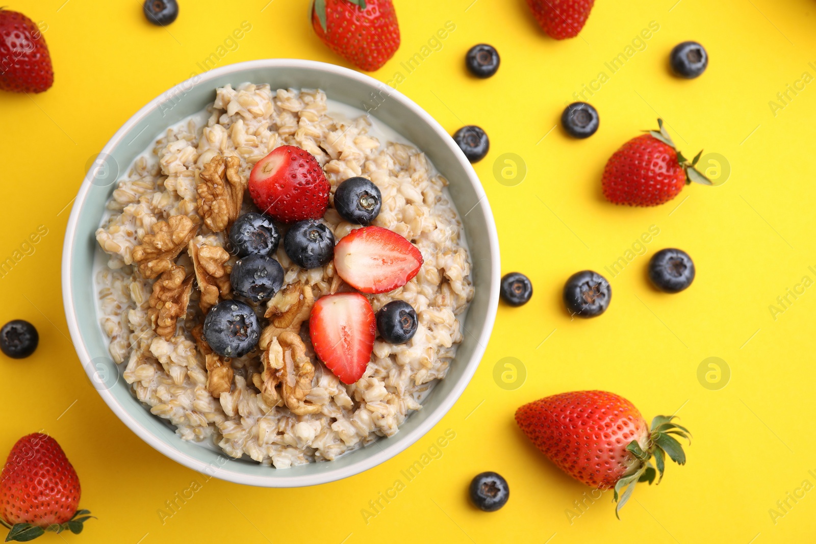 Photo of Tasty oatmeal with strawberries, blueberries and walnuts in bowl surrounded by ingredients on yellow background, flat lay