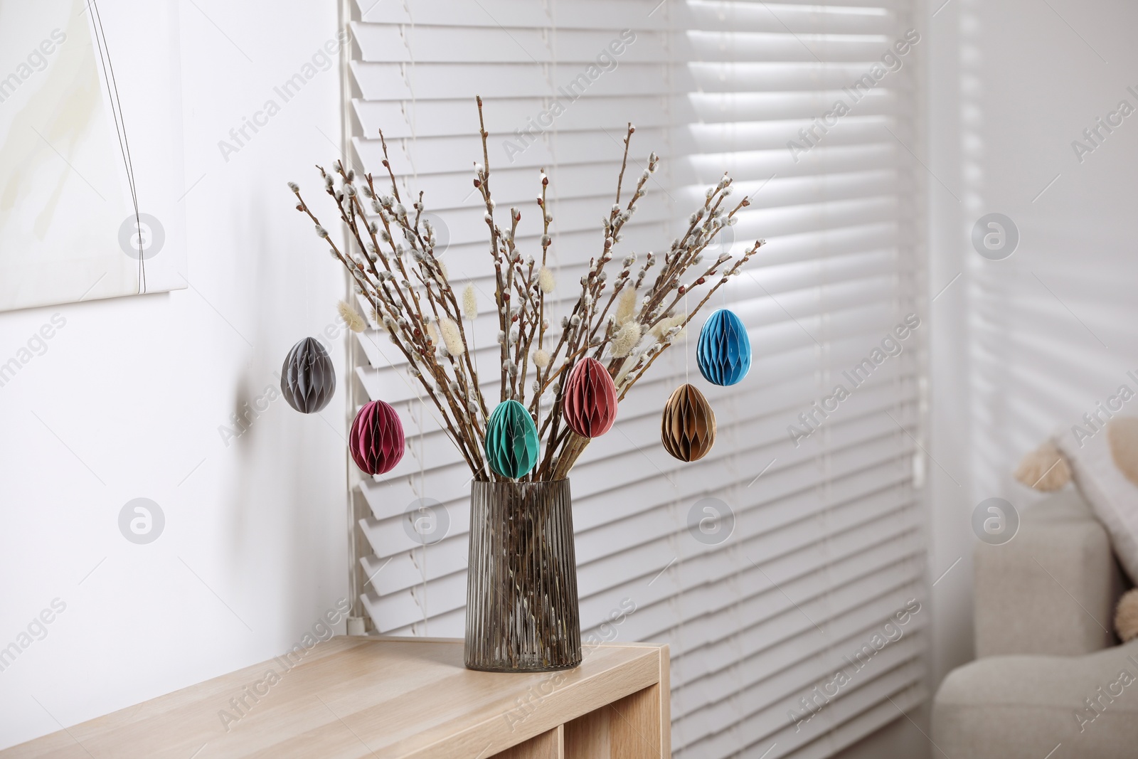 Photo of Beautiful pussy willow branches with paper eggs in vase on shelving unit at home. Easter decor